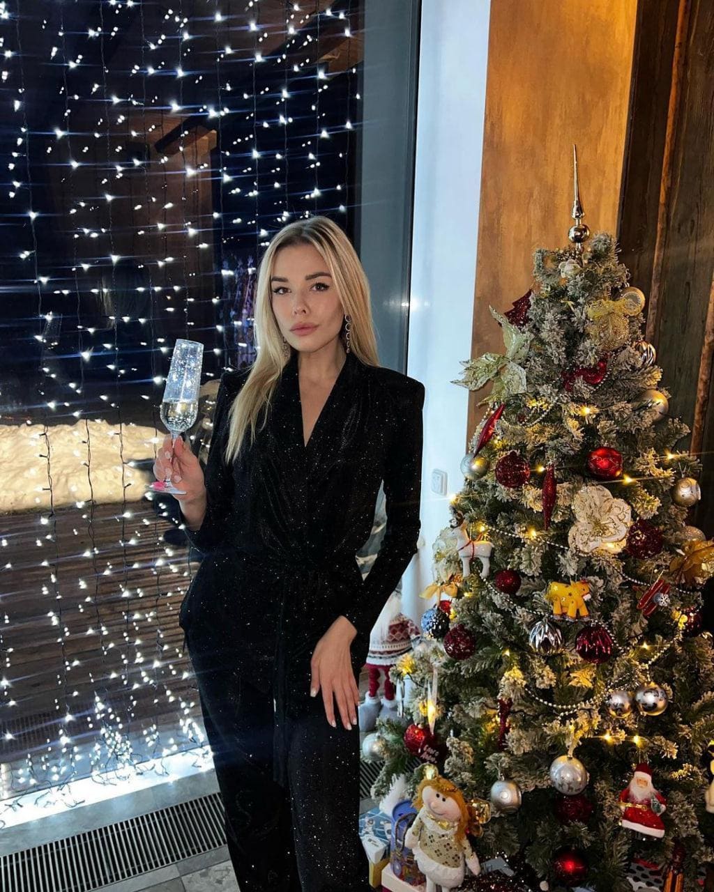 A blonde woman in a sparkly black wrap top and pants stands by a Christmas tree holding a sparkling drink, with string lights visible through a window.