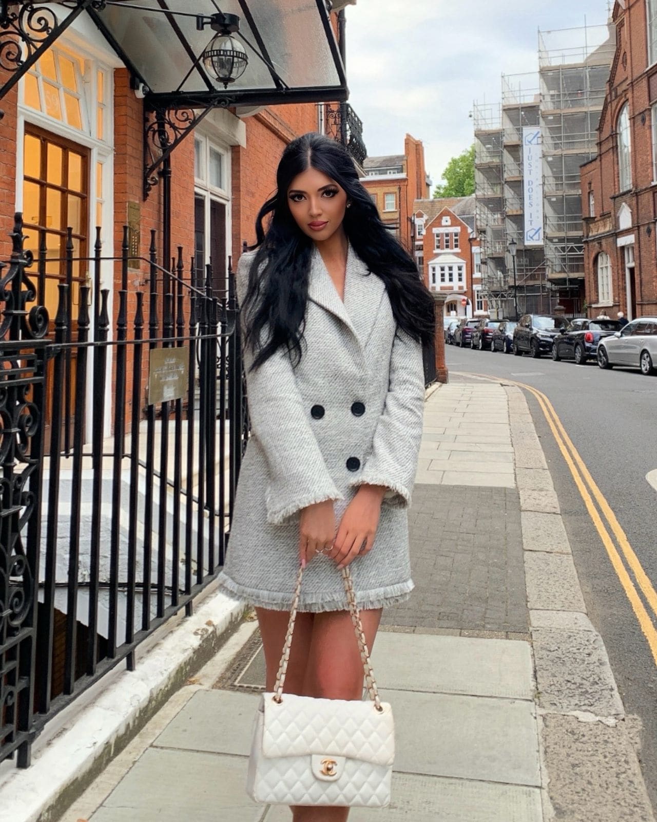 Autumn stands on a city sidewalk in a grey tweed coat dress, holding a white quilted handbag with brick buildings behind her.