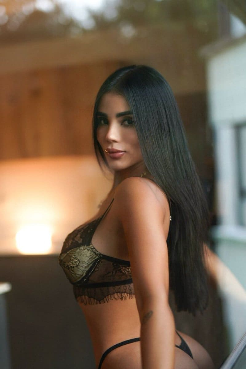 A woman in black and gold lace lingerie stands in profile facing the camera in a softly lit indoor setting.