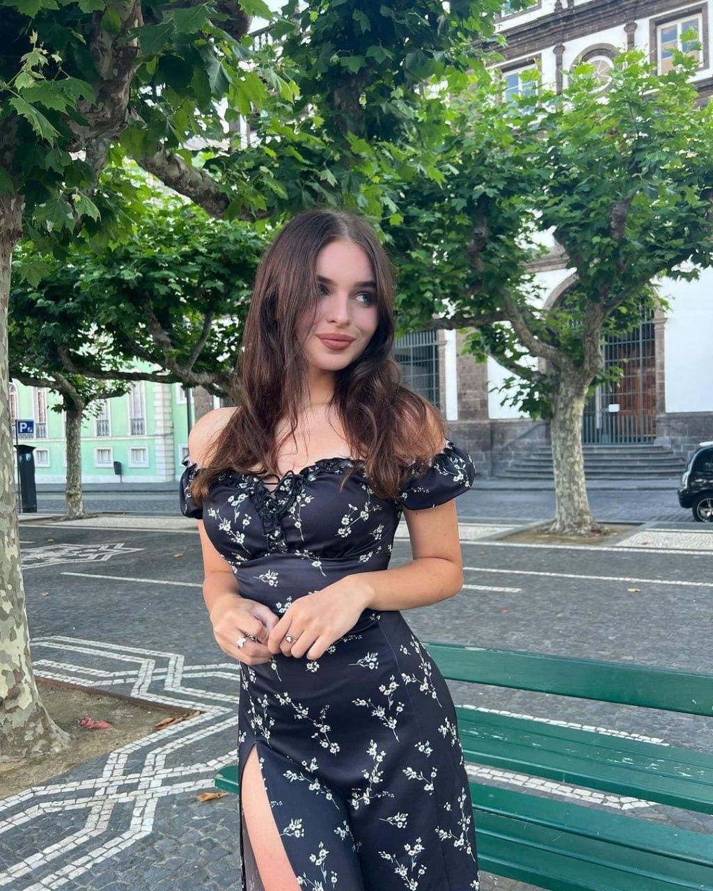 Natalie stands outdoors near a green bench in a black floral off-shoulder dress with a thigh-high slit and low neckline, trees behind.