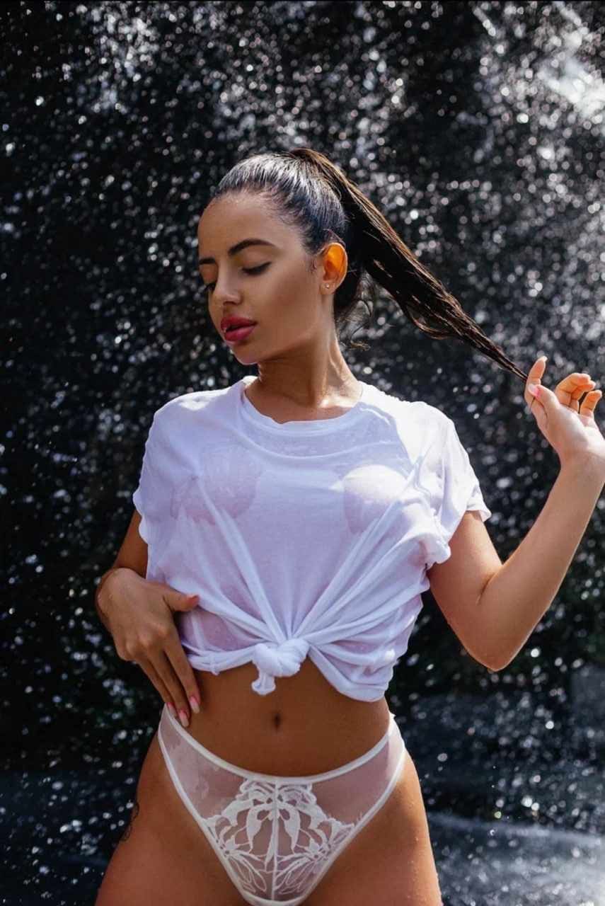 A woman with a wet white t-shirt and lace underwear stands against a dark background, holding her ponytail and resting a hand on her hip.