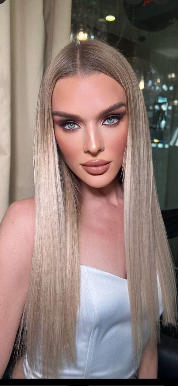 Zhenya sits facing the camera in a white strapless top, with long platinum-blonde hair, against a backdrop of curtains and a window.