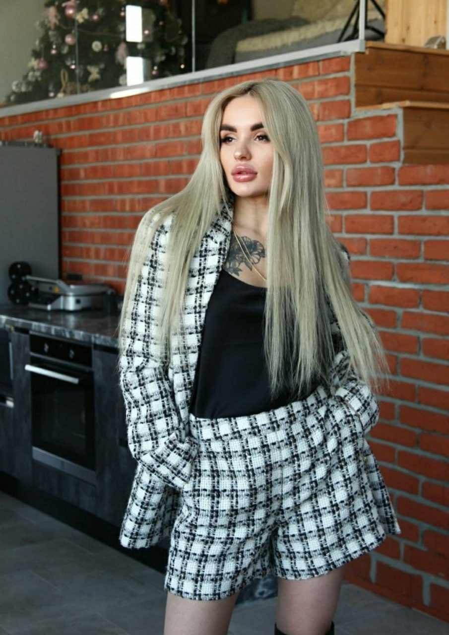 Woman with platinum hair in checkered blazer and shorts stands in kitchen with red brick wall, hands in pockets.