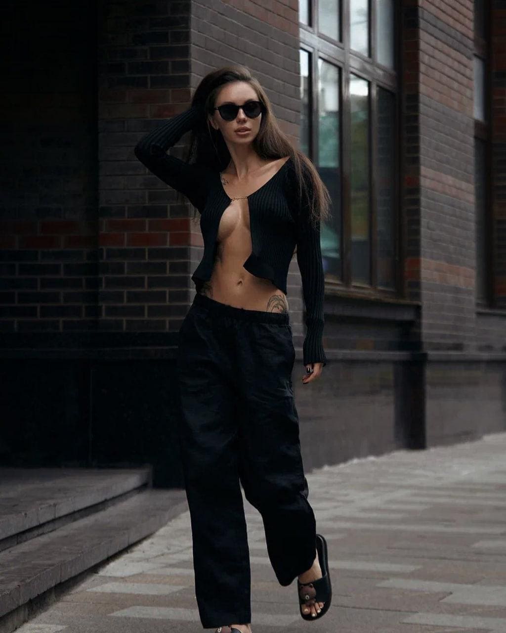 A woman in an open black cardigan and trousers stands on a street, touching her hair near a brick wall.