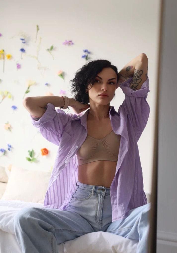 A woman sits on a bed with hands behind her head, wearing an open purple shirt over a beige bralette and jeans.