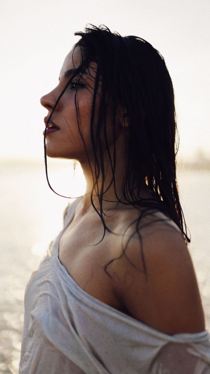 In profile against bright water, a woman wears a loose white top slipped off one shoulder, revealing her bare shoulder.