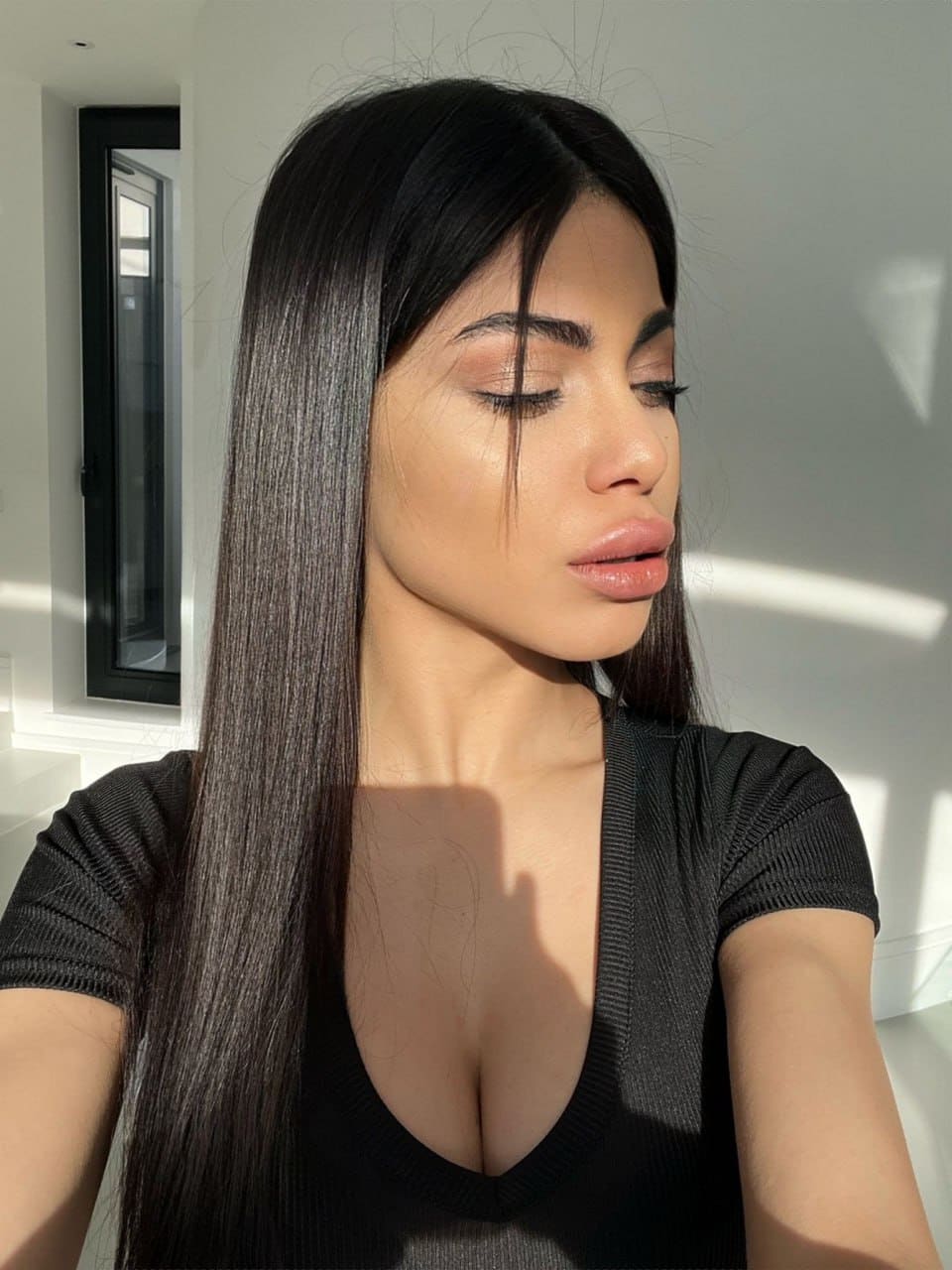 Cleopatra stands indoors near a window, eyes closed, wearing a black short-sleeved top with a deep V-neckline as sunlight casts shadows on the wall.