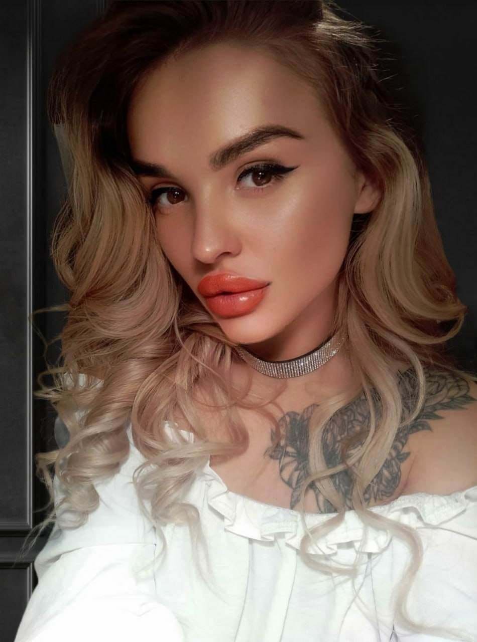 Angelica with long wavy blonde hair wears a white ruffled top and choker, showing a floral tattoo against a dark background.