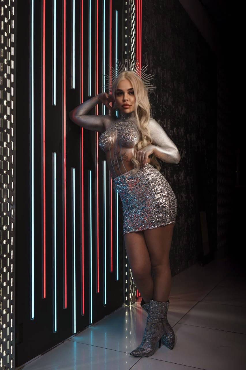 A blonde woman in a metallic headpiece, sheer sequined top, skirt, and boots poses with hand on hip against a dark wall with vertical neon strips.