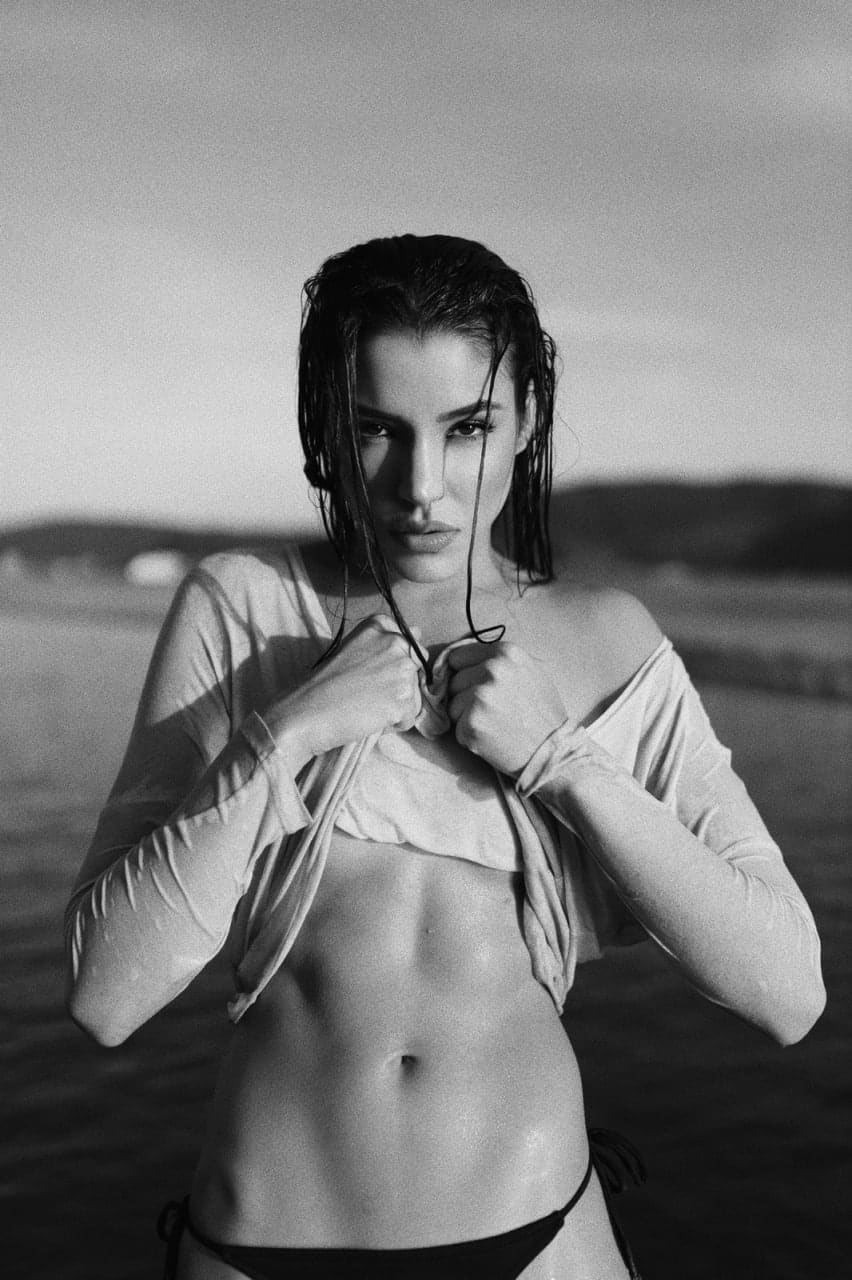 A woman with slicked-back wet hair pulls a light long-sleeve top down to expose her midriff in shallow water.