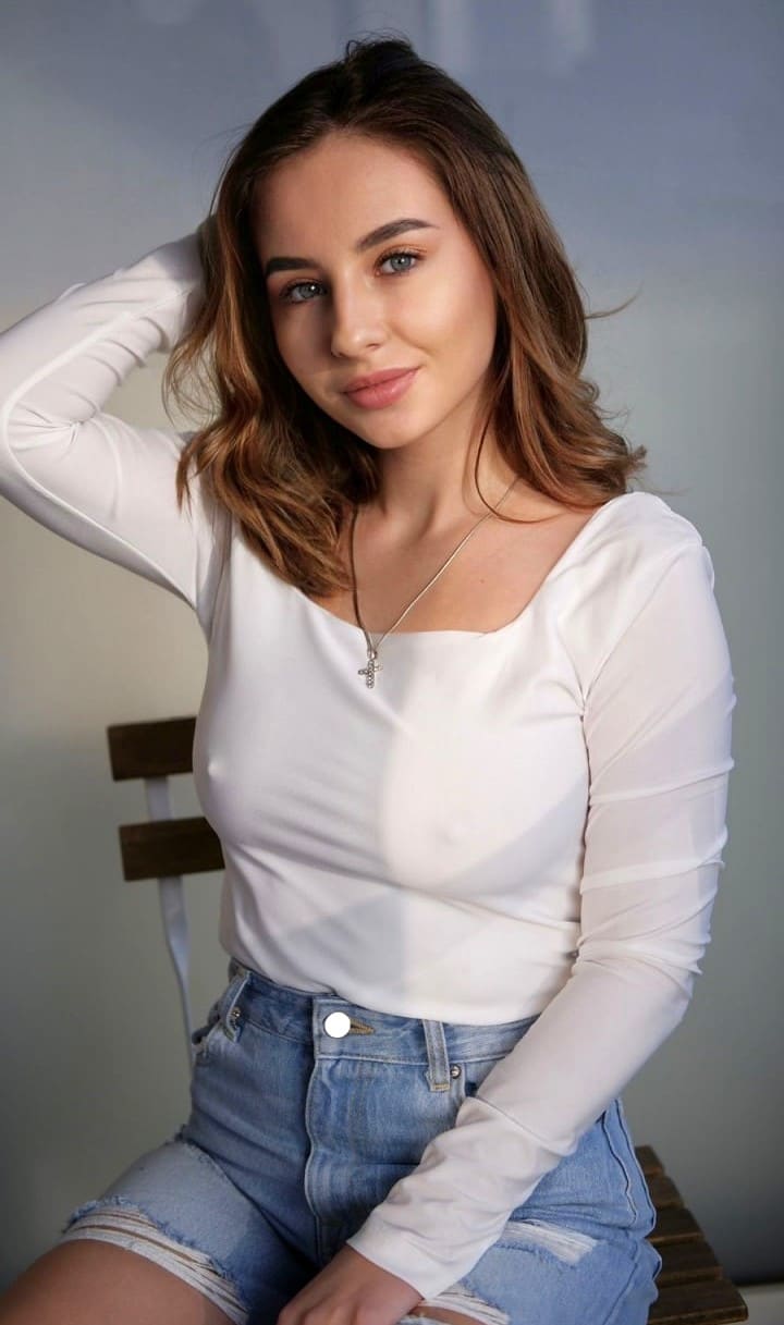 A woman in a white wide-neck top and denim shorts sits on a wooden chair, hand behind her head against a plain wall.