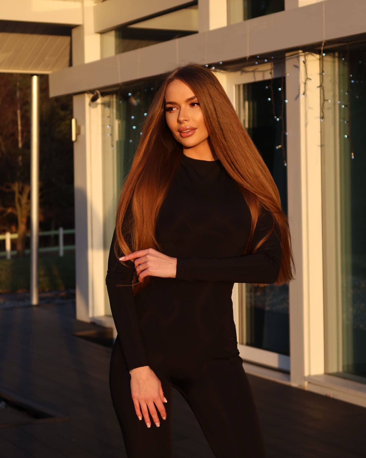 Standing on a wooden deck before a modern glass building, she wears a fitted black long-sleeve top and pants, hand near her waist.