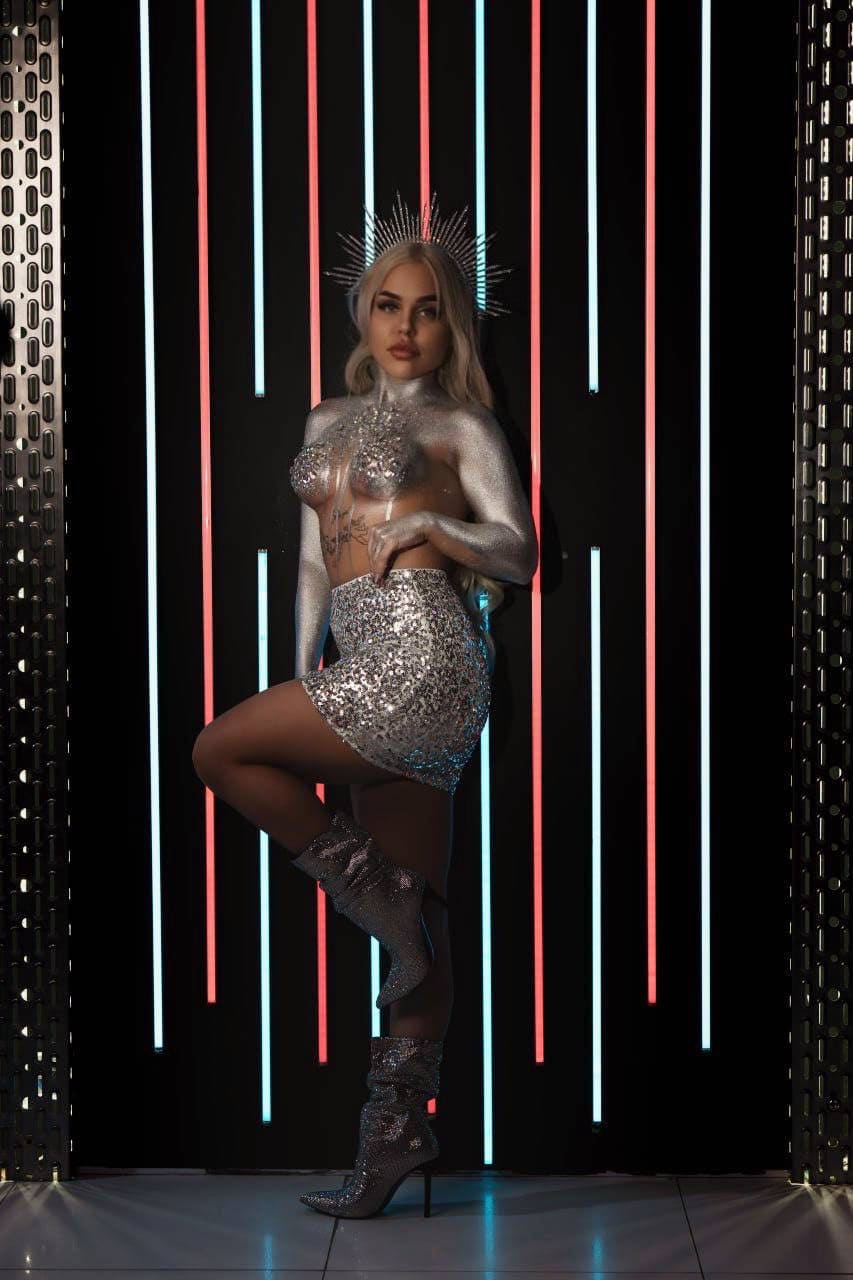 A blonde woman with silver body paint and a spiky headpiece poses in a sequined skirt, sheer top, and tall silver boots against neon lights.