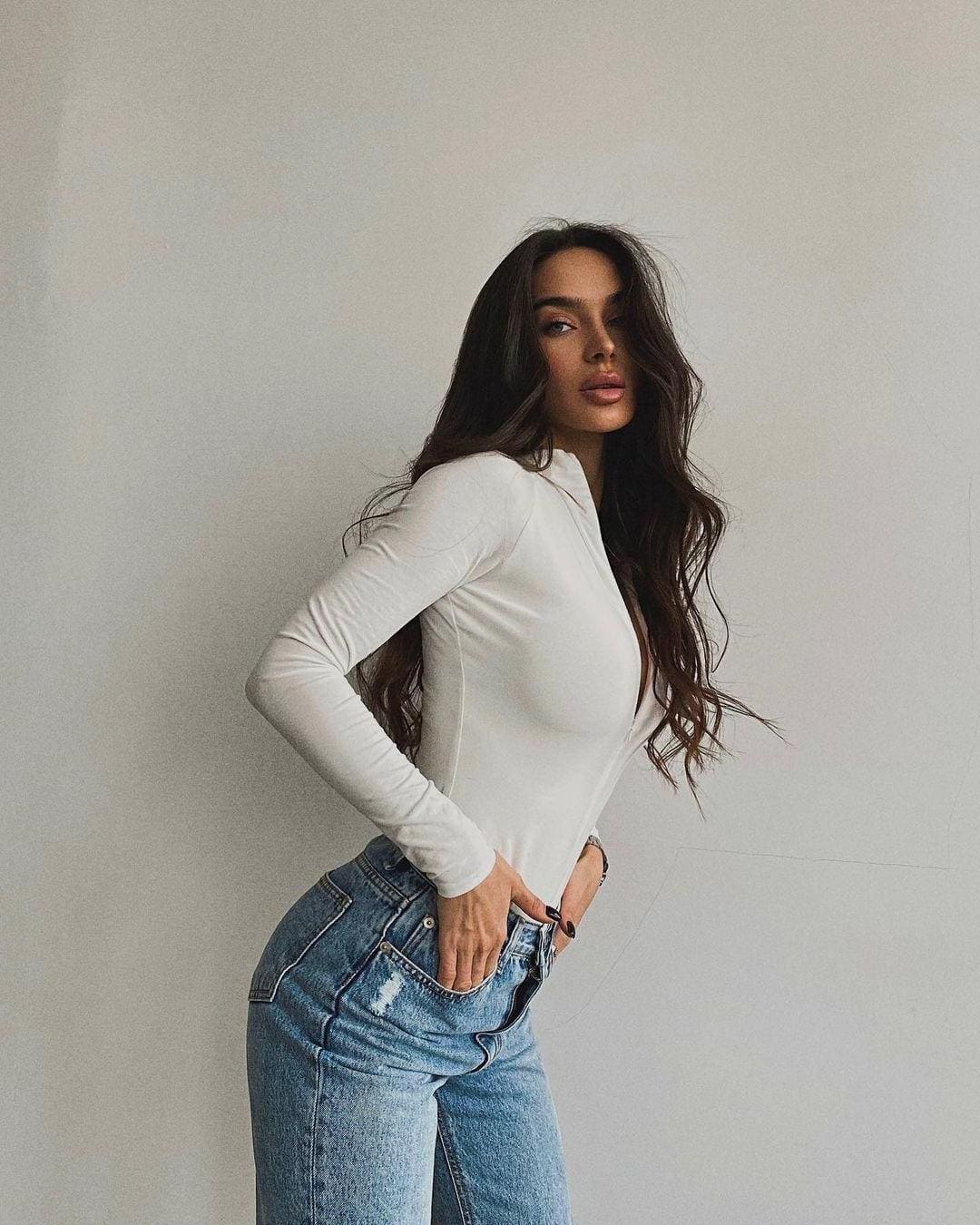 A woman with long wavy hair stands in profile against a light wall, wearing a white long-sleeve top and distressed jeans.