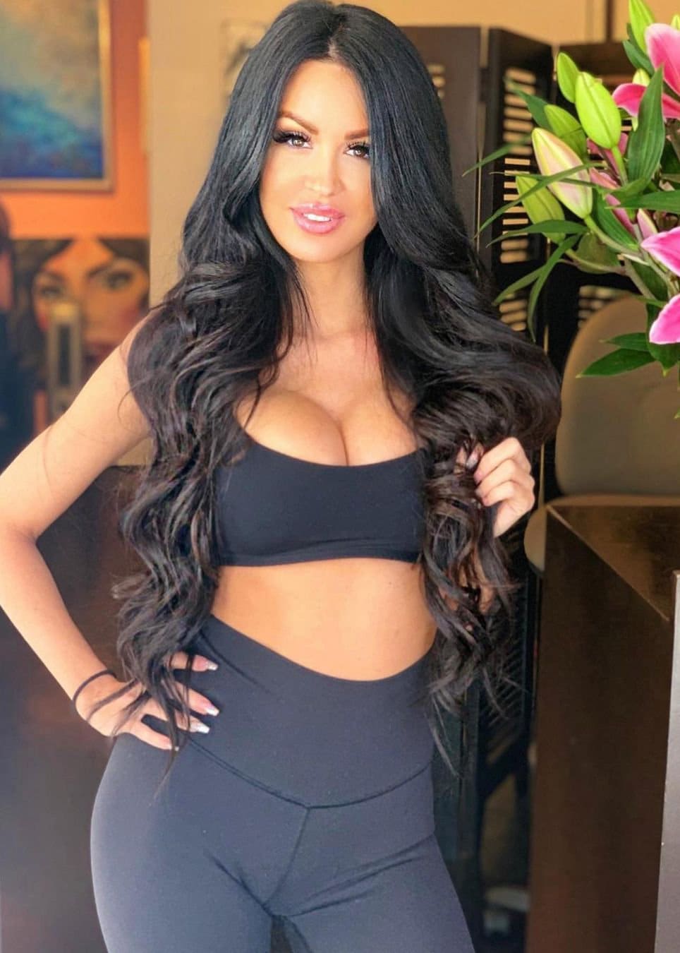 Jamie stands indoors in a black strapless crop top and leggings, one hand on her hip, holding hair, with lilies and art behind.