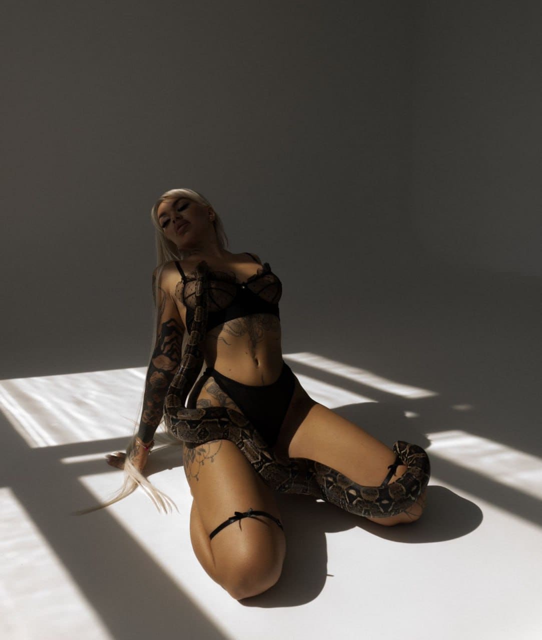 A tattooed blonde woman kneels in black lace lingerie with a garter strap, a large snake coiled around her torso in a dim room.