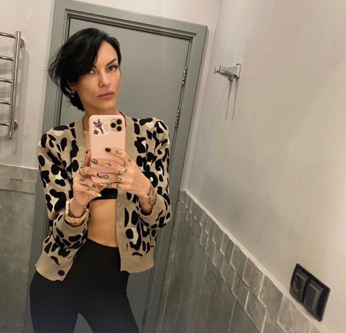 A woman takes a bathroom mirror selfie in an animal-print cardigan over a black bra and leggings, holding a pink phone.