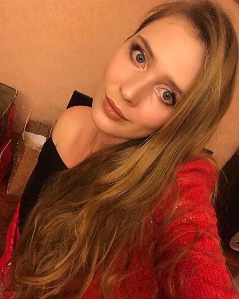 Close-up selfie of a blonde woman in a red sequined top over a black off-shoulder garment, against a warm indoor wall with boxes.