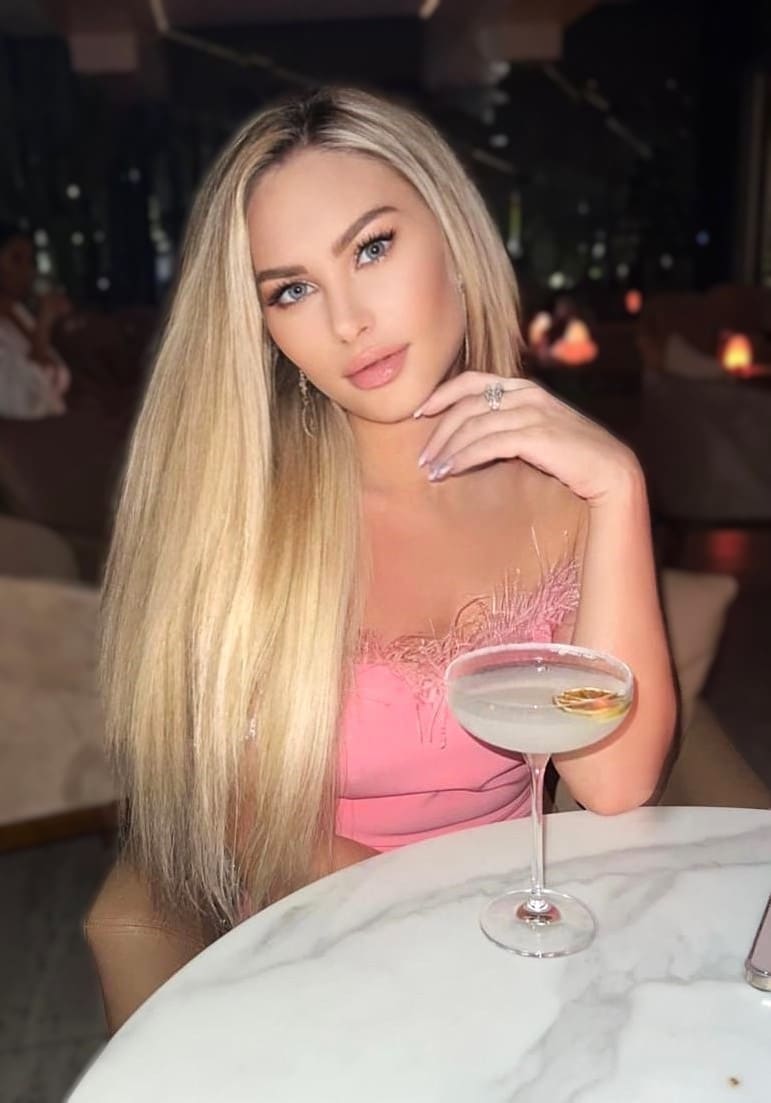 Woman in a pink lace top sits at a marble table in a dim lounge, hand near chin, with a cocktail glass in front.