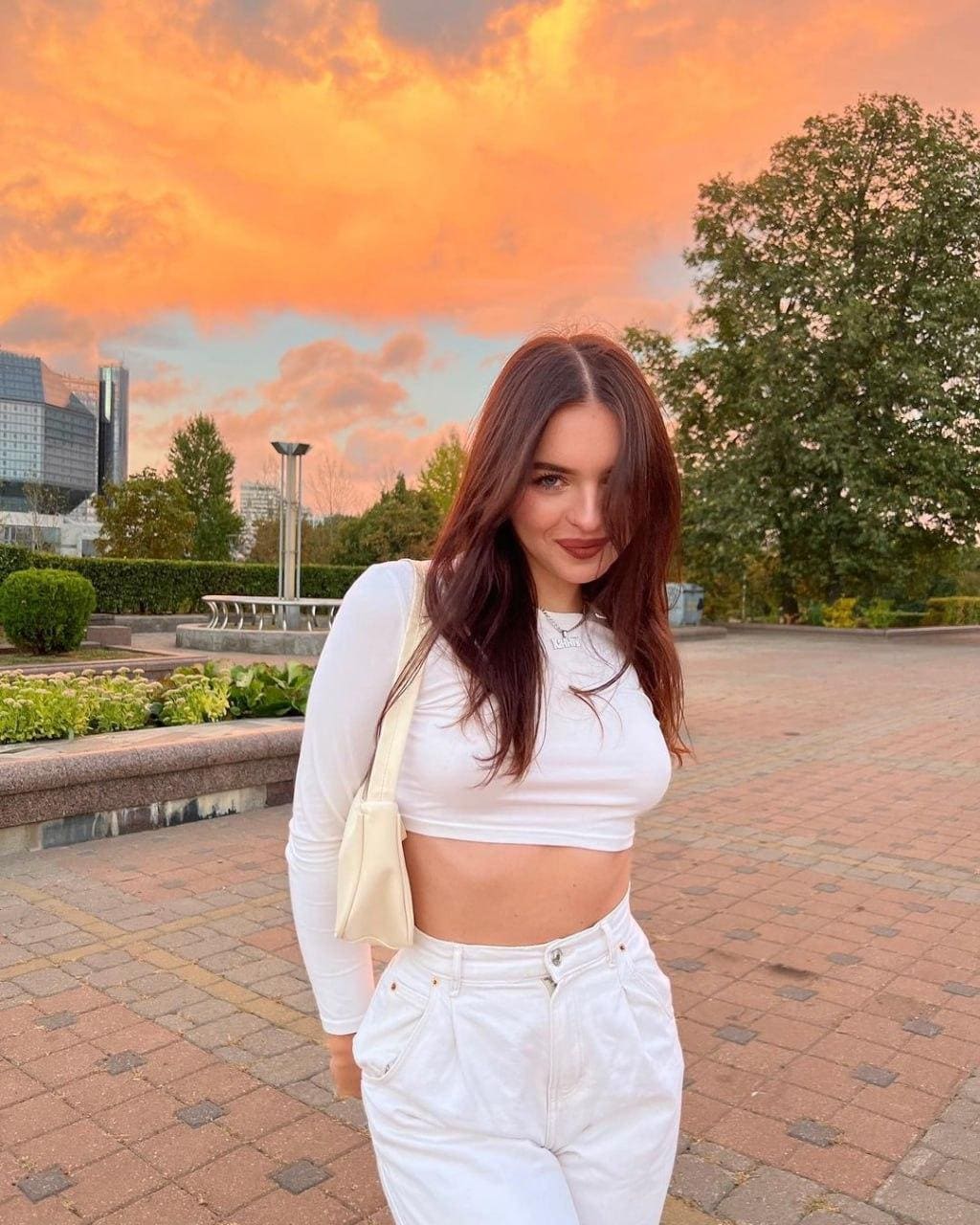 A woman in a white crop top and jeans stands on a brick plaza at sunset, holding a cream bag against an orange sky and distant buildings.