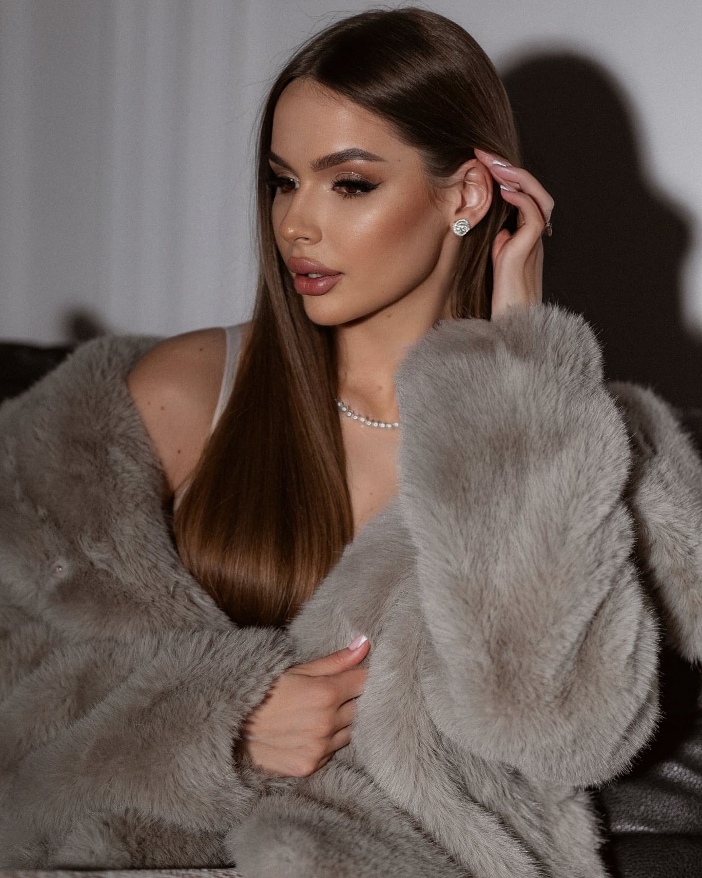 Aurora sits indoors in a grey fur coat over a light top, one hand at her ear and the other on her chest against a plain backdrop.