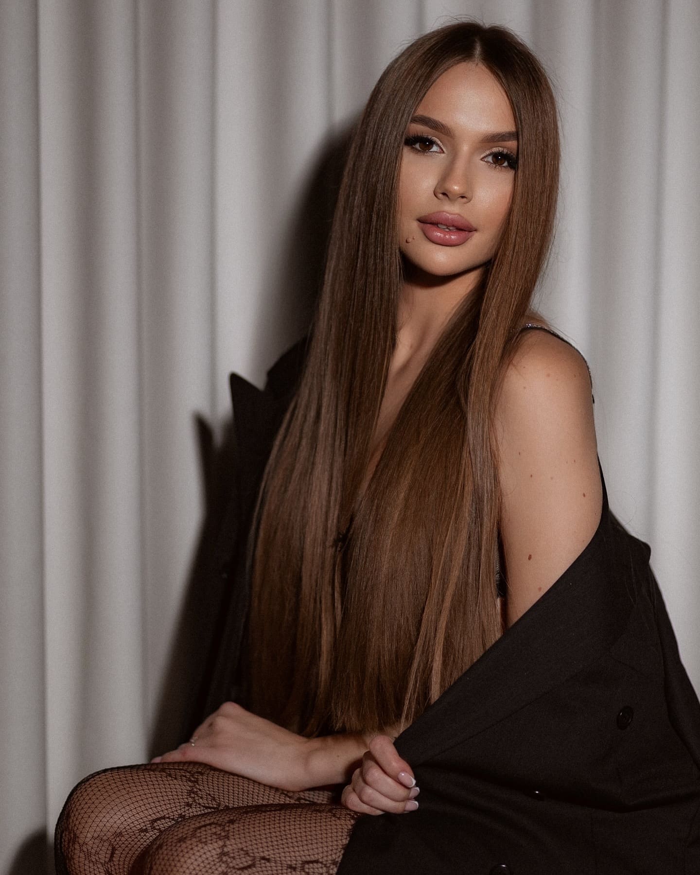 In front of a white curtain, she sits in a black sleeveless top and draped blazer, fishnets visible on her crossed legs.