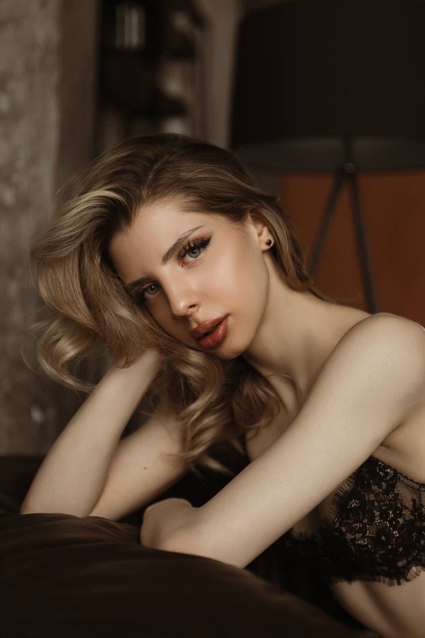 A woman leans forward with crossed arms on a dark surface, wearing a black lace strapless top in dim lighting.