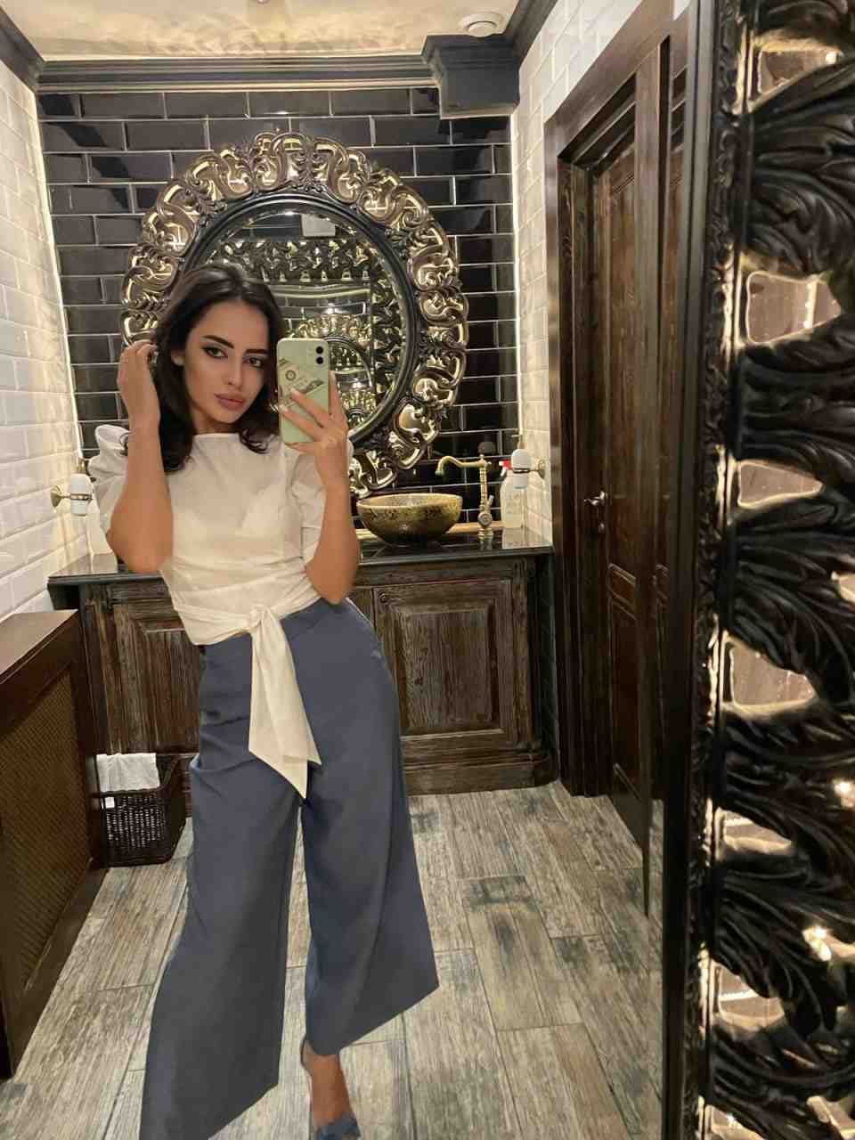 A woman in a tied white blouse and grey trousers takes a bathroom mirror selfie, one hand touching her hair.