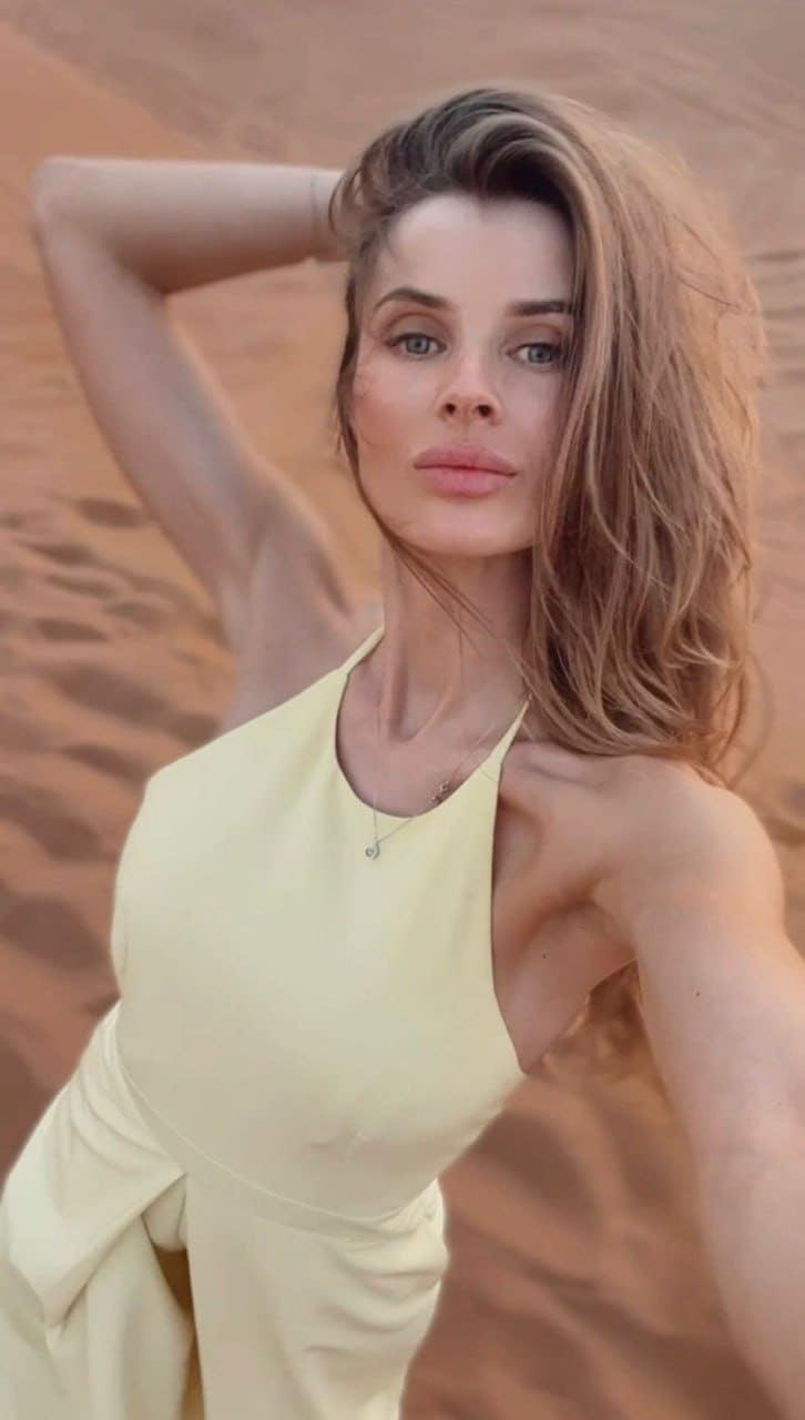 A woman in a pale yellow dress stands in a desert with one arm raised behind her head, wearing a thin necklace.