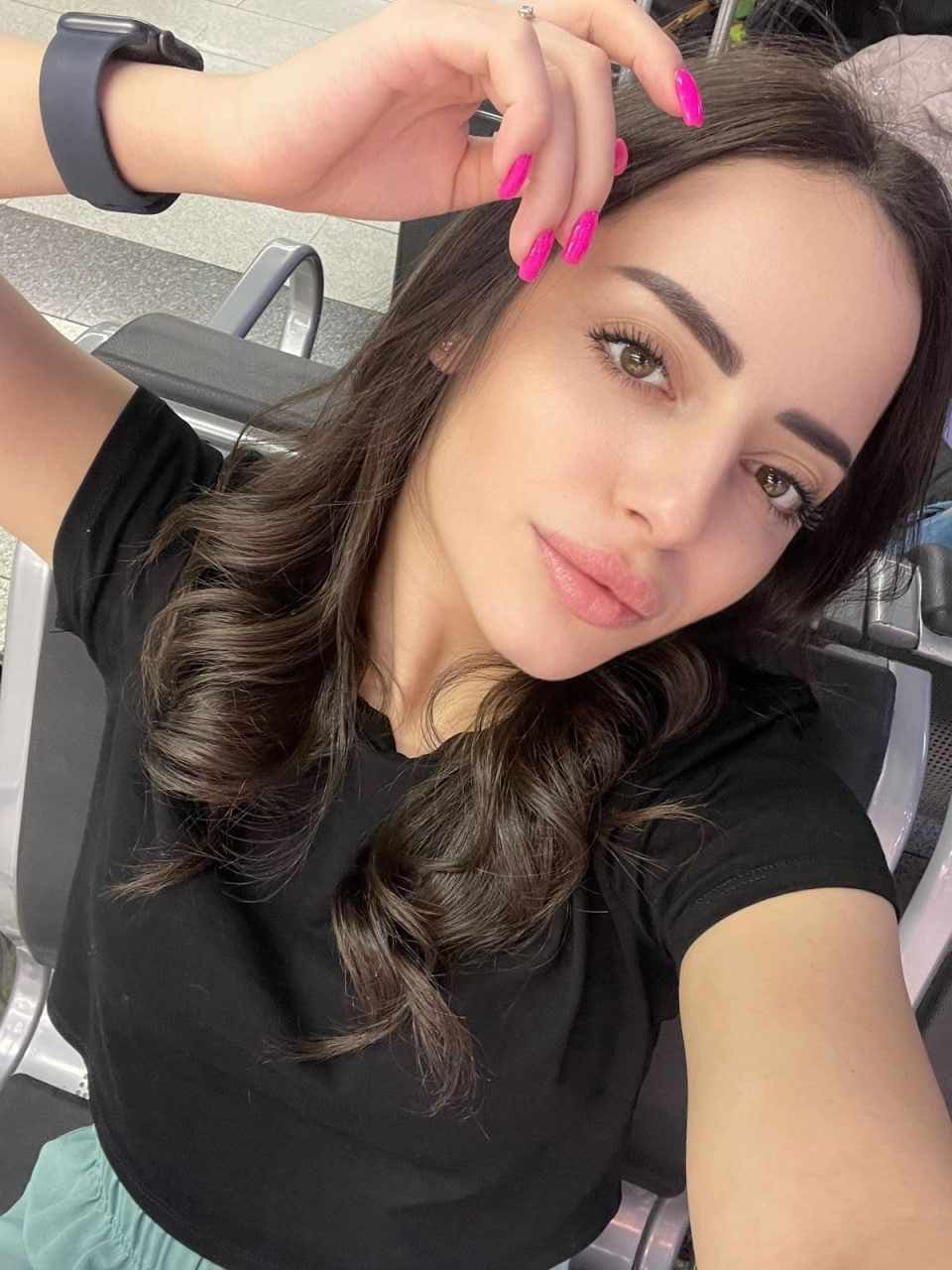 A close-up selfie shows a woman with pink nails and a black t-shirt seated in a chair with metal armrests.