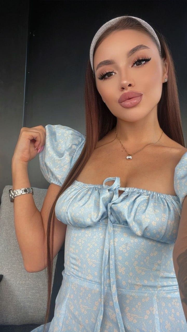 Angelina poses in a light blue floral off-shoulder dress with puffed sleeves and a headband, wearing a necklace and watch, against a dark background.