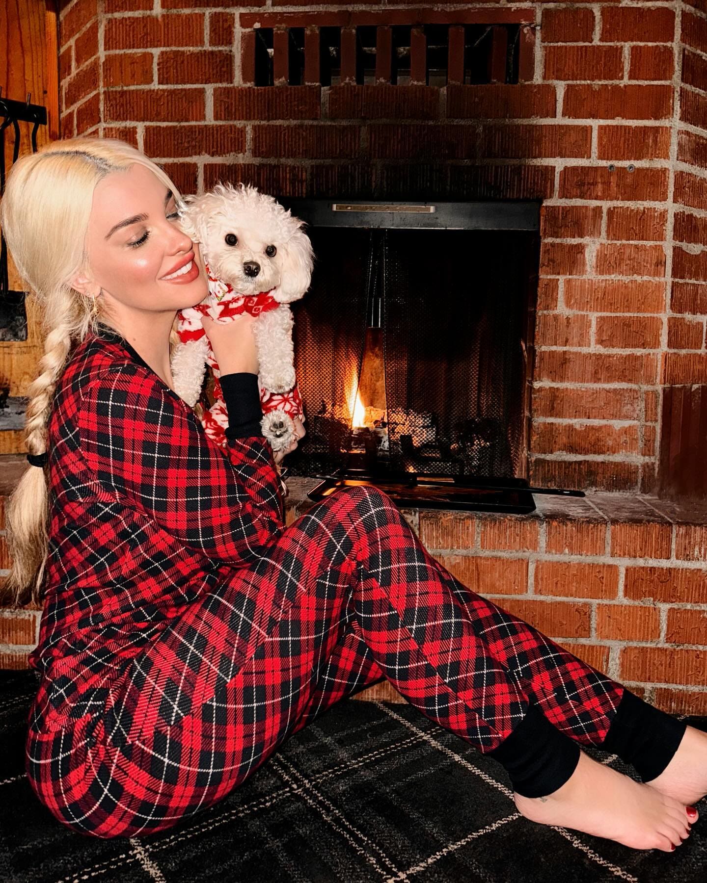 A blonde woman in red plaid pajamas sits barefoot on a rug by a fireplace, holding a small white dog in matching attire.