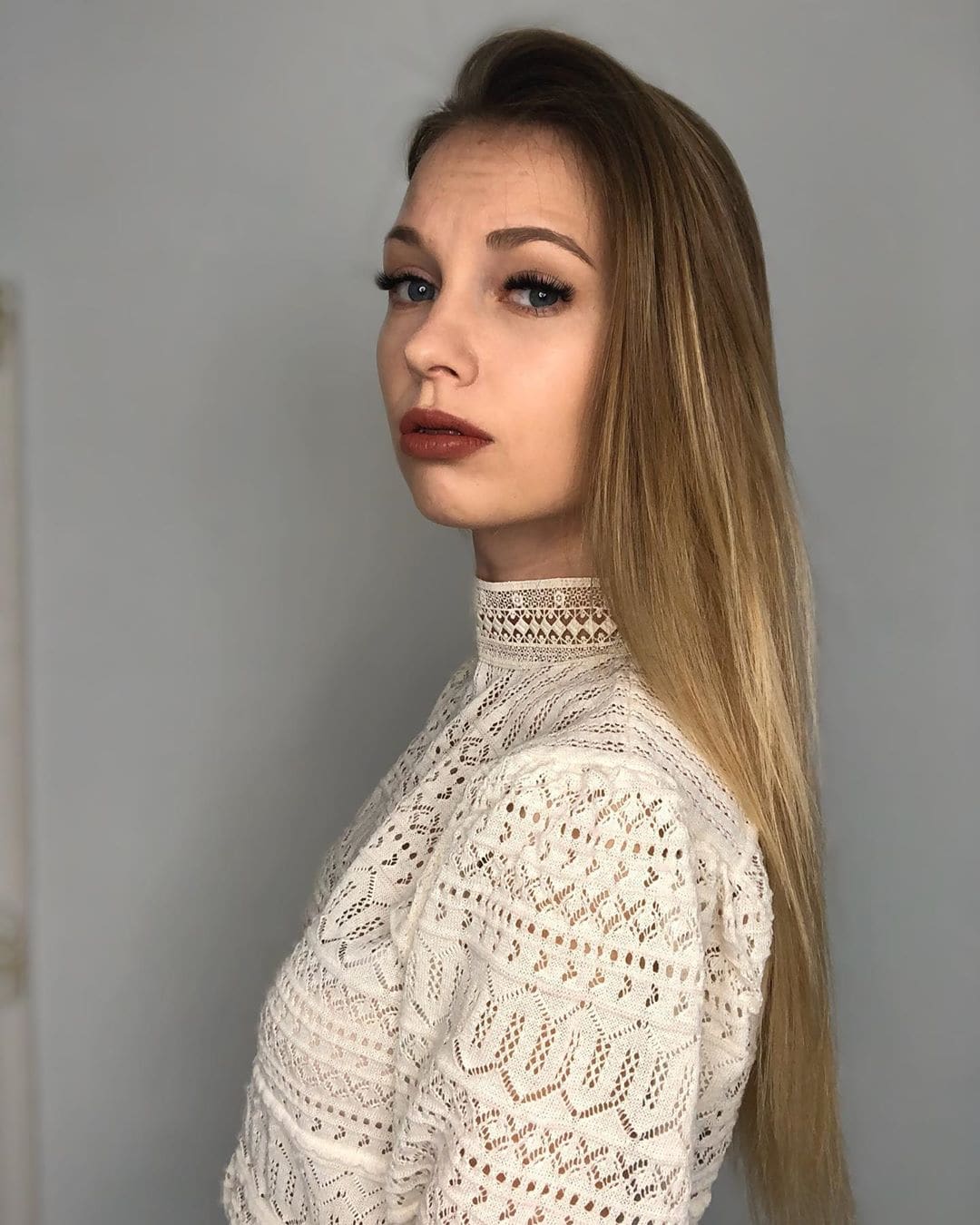 Karoline in a white high-neck lace top, three-quarter profile against a plain gray wall.