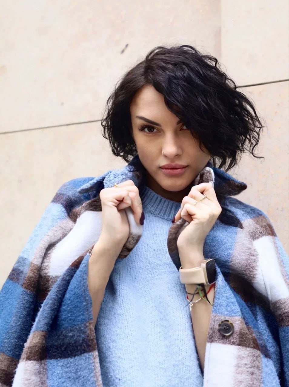 A woman stands against a stone wall in a blue knit sweater and plaid coat, holding the collar with both hands.