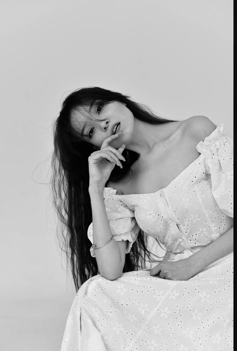 A woman in a light off-the-shoulder eyelet dress sits in a studio, hand near her chin.