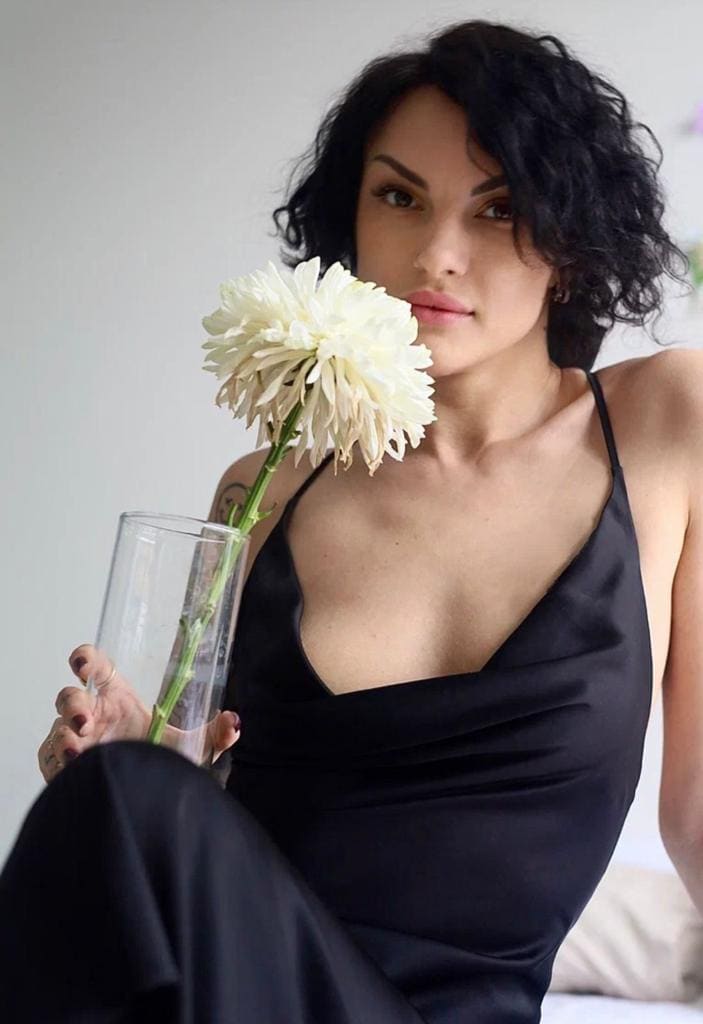Risha sits holding a white dahlia in a glass vase, wearing a black satin slip dress against a light wall.