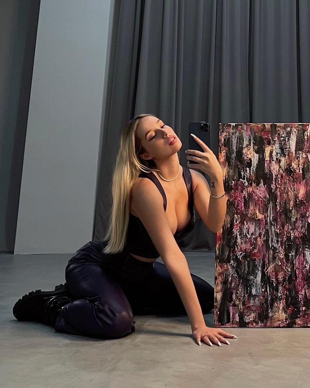 A blonde woman kneels on a concrete floor in a black top and dark pants, taking a mirror selfie with a phone near an abstract painting.