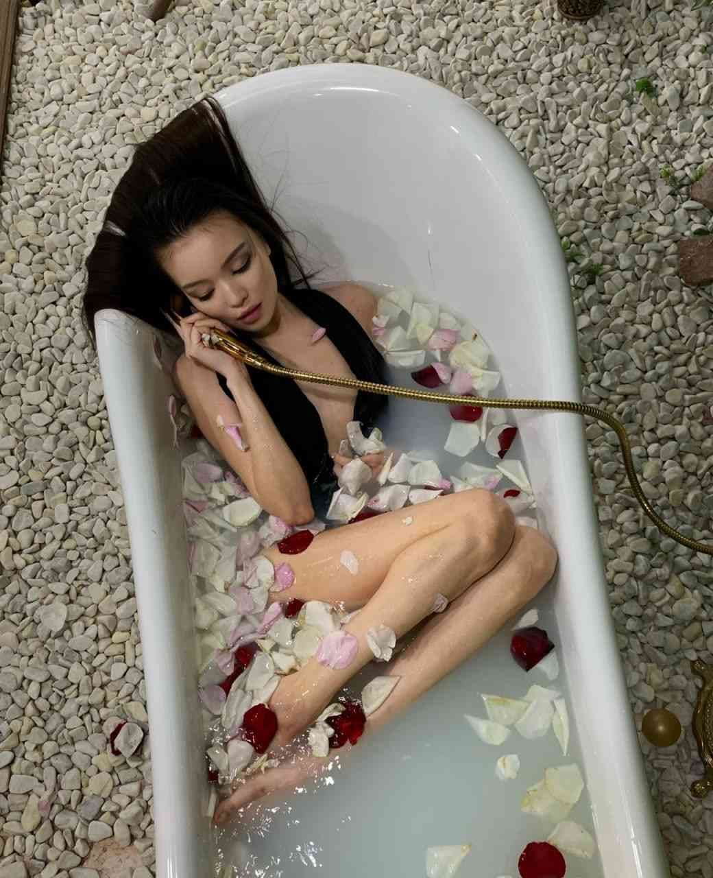 A woman in a black swimsuit lies in a bathtub with rose petals, holding a gold shower head.