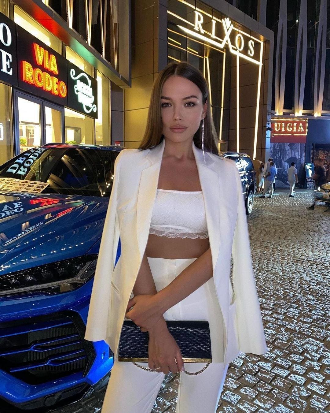 A woman in a white blazer, lace crop top, and pants holds a black clutch while standing on a cobblestone street at night near a blue car.