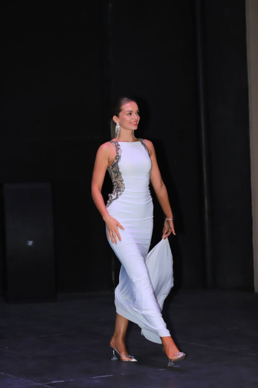 Walking on a dark stage, a woman in a white lace gown lifts the hem with one hand.