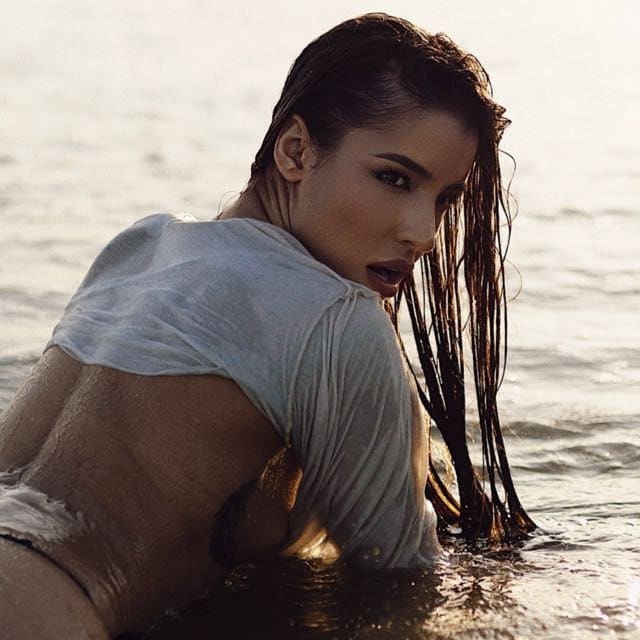 Partially submerged, a woman in a soaked white top looks over her shoulder, wet hair clinging to her back.