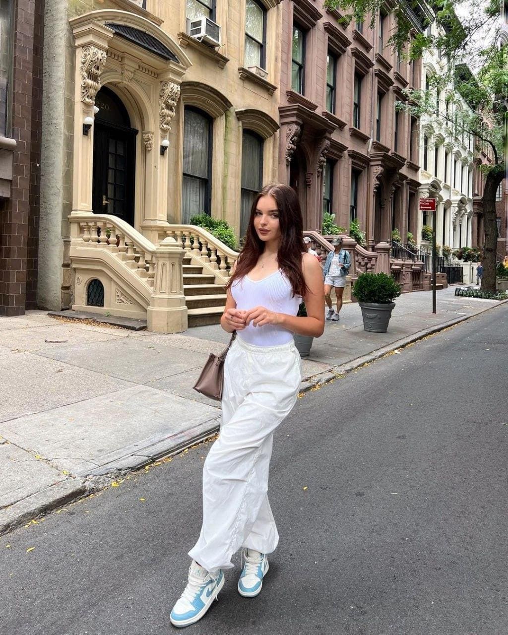 A woman in a white tank top and trousers stands on a city street before brownstones, holding a brown bag and wearing blue sneakers.
