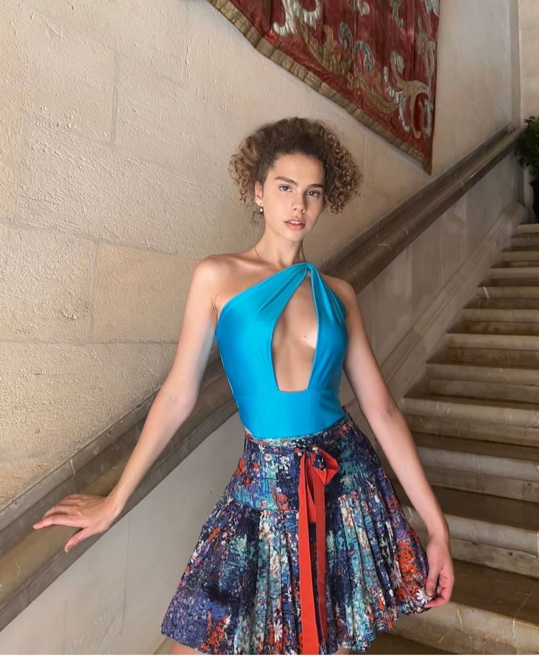 Zoey stands on a stone staircase in a turquoise one-shoulder top and floral pleated skirt, resting her hand on the railing.