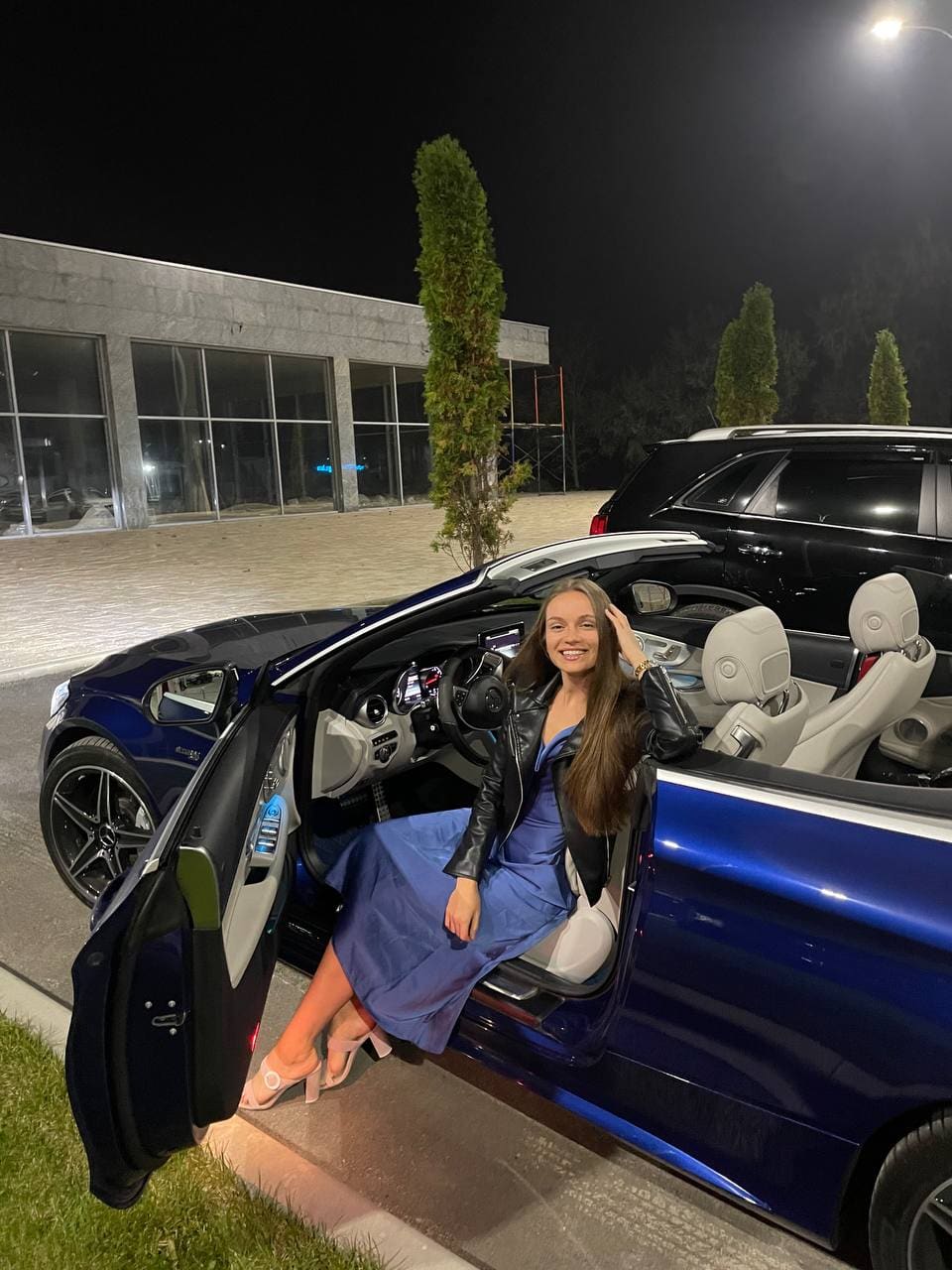 At night, a woman in a blue dress and leather jacket sits in the door of a blue convertible car.