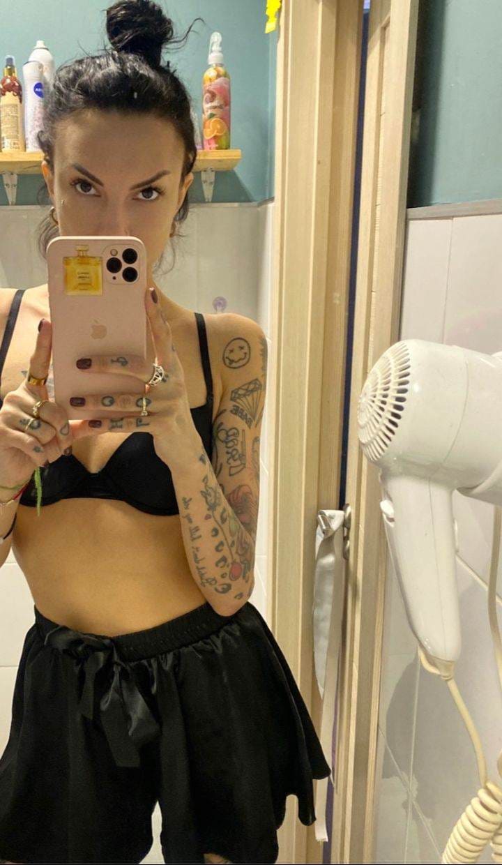 A woman with a high bun takes a bathroom mirror selfie in a black bra and drawstring shorts, holding a pink phone.