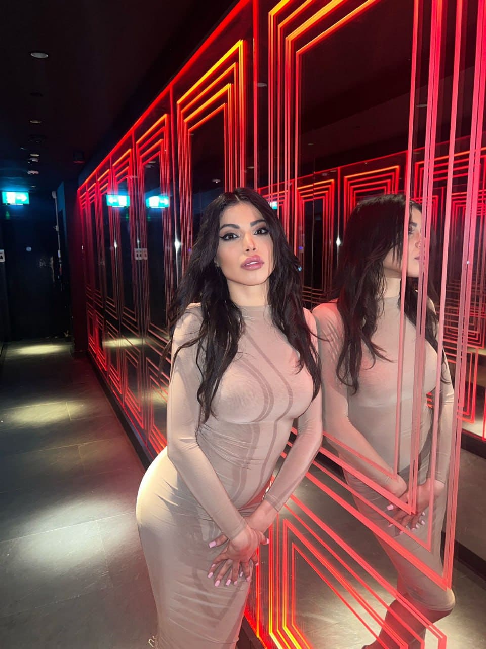 A woman stands in a hallway with red neon panels, wearing a long-sleeved beige dress that is semi-sheer across the chest, hands clasped in front.
