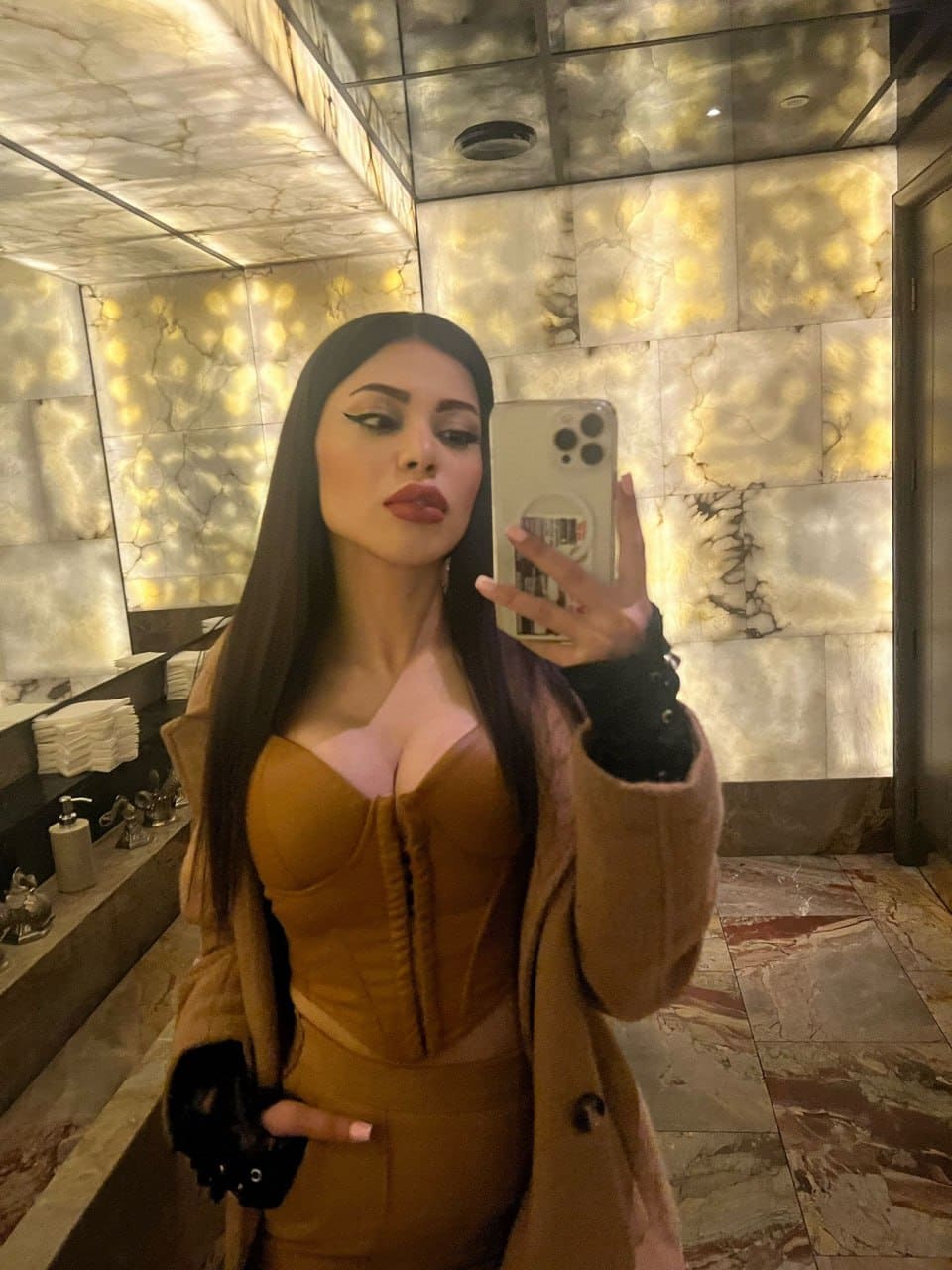 A woman takes a mirror selfie in a marble-walled room, wearing a brown corset under an open tan coat and black fingerless gloves.