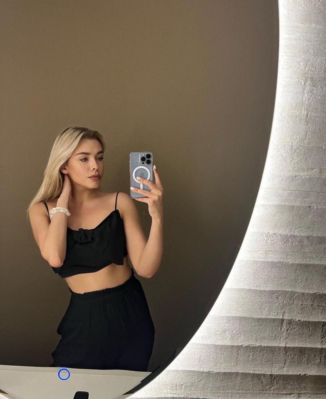 A blonde woman takes a mirror selfie in a black crop top and high-waisted bottoms, holding a phone and touching her neck against a plain wall.