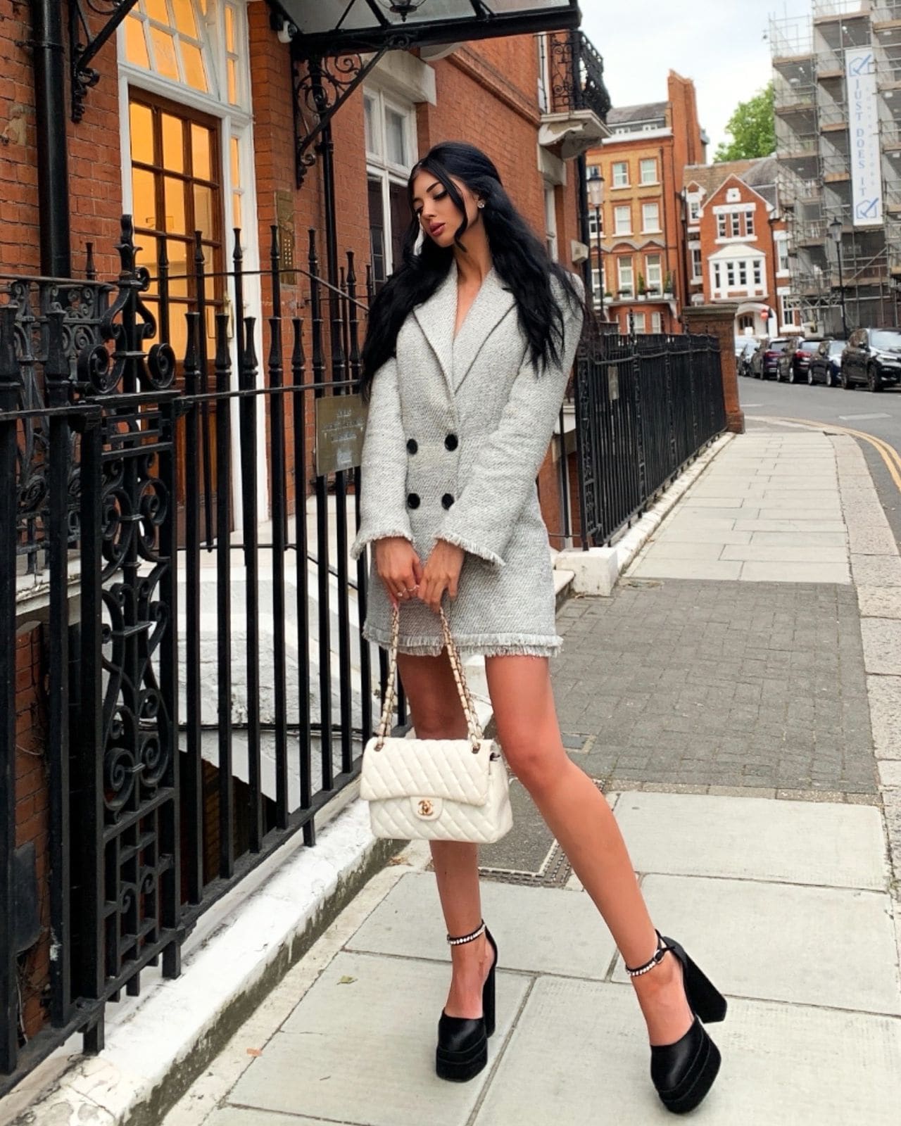 Woman in a grey tweed mini dress and platform heels holds a white handbag on a sidewalk near a black iron fence.