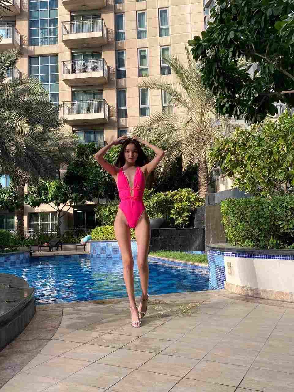 A woman in a pink one-piece swimsuit stands by a pool with hands raised, palms and building behind.
